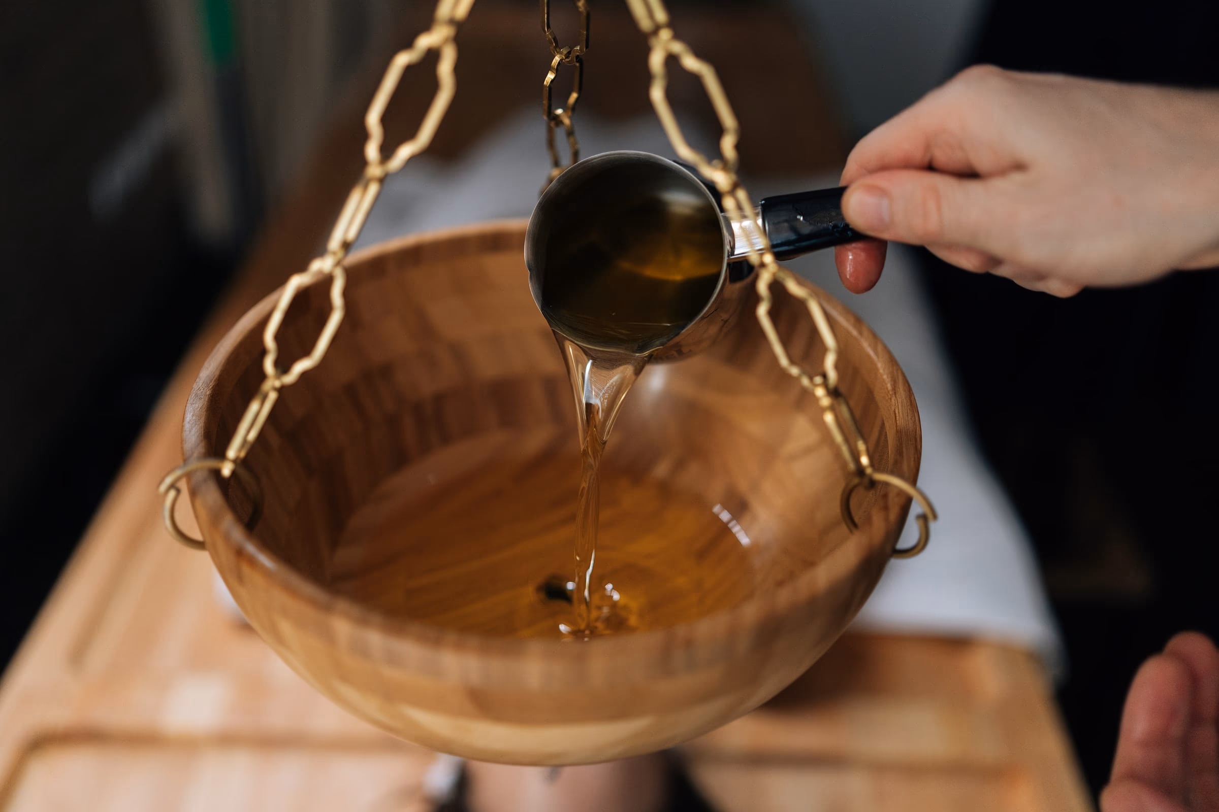 Warm herbal oil being poured from a small pan into a suspended wooden dhara vessel before a treatment